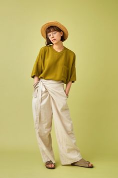 a woman standing in front of a green background wearing a straw hat and linen pants