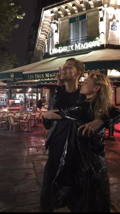 two people standing on the sidewalk in front of a restaurant at night with their arms around each other