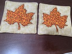 two quilted coasters with leaves on them sitting on a wooden table next to a knife