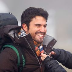 a man smiles as he adjusts his jacket on the back of a motorcycle with a backpack behind him