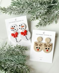 two christmas themed earrings on top of a card next to some evergreen branches and snowflakes