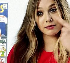 a woman in a red shirt is holding her hand up to her ear and looking at the camera