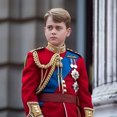 a young boy dressed in red and blue is standing on the balcony of buckingham palace