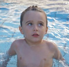 a young boy swimming in a pool with no shirt on, looking at the camera