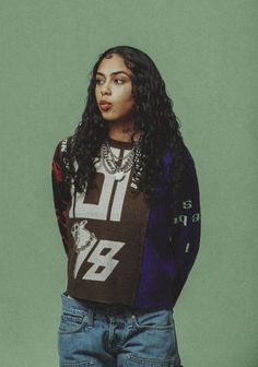 a woman with long curly hair wearing a sweater and jeans standing in front of a green background