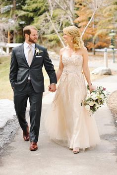 a bride and groom holding hands walking down the street in their wedding attire, dressed in grey tuxedos