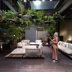 a woman is standing in front of some couches and tables with plants hanging from the ceiling