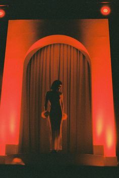 a woman standing in front of a stage with red lights on the walls and curtains behind her