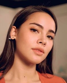 a woman with long hair wearing an orange top and earrings, looking at the camera