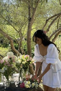 a woman in a white dress arranging flowers on a table with vases and trees behind her