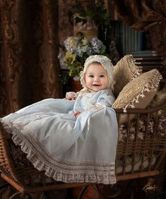 a baby in a white dress is sitting on a wicker bed with pillows and flowers