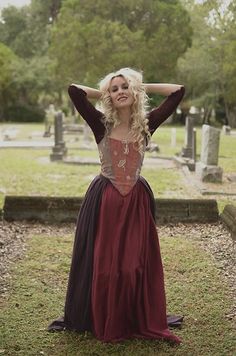 a woman with blonde hair wearing a long dress in front of a grave yard and trees