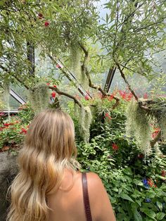 a woman standing in front of some flowers and trees with long blonde hair looking down at the ground