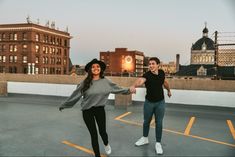 two people holding hands and walking across an empty parking lot with buildings in the background