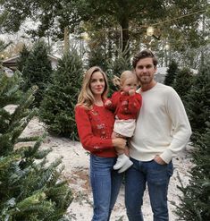 a man and woman standing next to each other in front of some christmas trees with one holding a baby