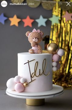 a teddy bear sitting on top of a white cake with gold letters and balloons around it