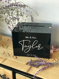 a wooden table topped with a black box filled with purple flowers and a couple's initials on it