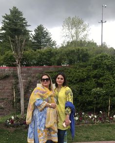 two women standing next to each other in front of a brick wall and green grass