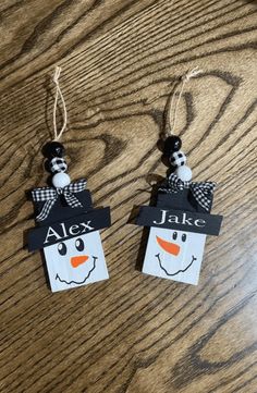 two snowmen with name tags hanging from strings on a wooden table, one is wearing a black and white bow