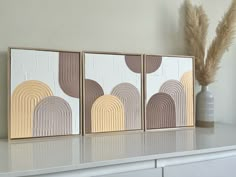 three framed art pieces sitting on top of a white shelf next to a vase with dried grass in it