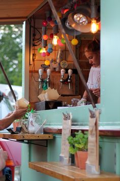 a woman is preparing food at an outdoor stand with lights hanging from the ceiling and potted plants on the counter