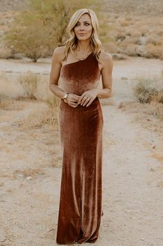 a woman standing in the desert wearing a brown velvet dress with one shoulder draped over her shoulders