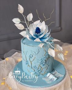 a blue cake with white flowers and leaves on top is sitting on a lace tablecloth