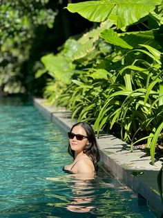 a woman wearing sunglasses is in the water near some plants and bushes, with her back turned to the camera