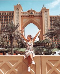 a woman sitting on top of a stone wall in front of a large building with palm trees