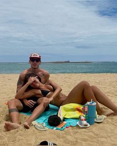 two people are sitting on the beach and one is holding his arm around the other