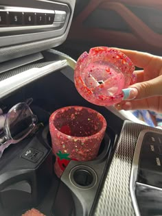 a person is holding a pink cup in their hand next to other items on the car dashboard