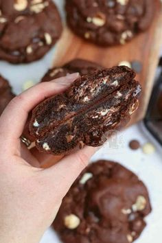 a person holding a chocolate cookie in front of some cookies on a cutting board with one bite taken out