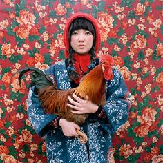 a woman holding a rooster in front of a red floral wallpaper with flowers on it