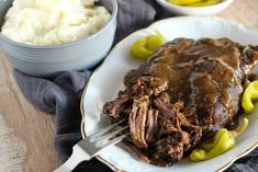 a white plate topped with meat covered in gravy next to two bowls of mashed potatoes