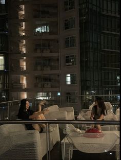 two women sitting on white couches in front of a tall building at night,