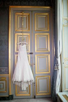 a wedding dress hanging in front of an open door to a room with gold trimmings