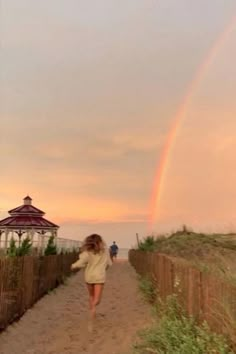 a woman walking down a dirt road next to a fence and a rainbow in the sky