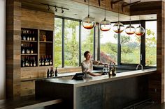 a woman sitting at a bar in front of large windows with wine bottles on it