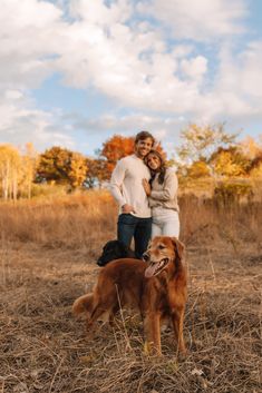 a man and woman standing next to a brown dog on top of a dry grass field