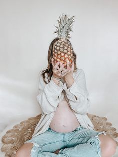 a pregnant woman with a pineapple on her head sitting in front of a white background