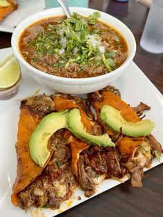 a white plate topped with food next to a bowl of soup and an avocado