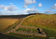 an aerial view of a grassy hill with a stone wall in the foreground and clouds in the background