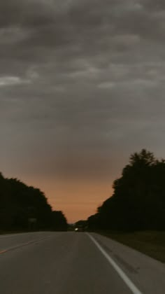 an empty road with trees on both sides and dark clouds in the sky above it