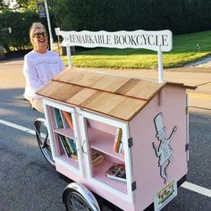 a woman standing next to a pink book cart with books on the side and a sign that says remarkable bookcycle