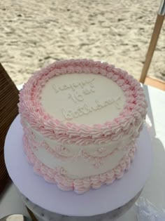 a pink and white cake sitting on top of a table next to the ocean with happy birthday written on it