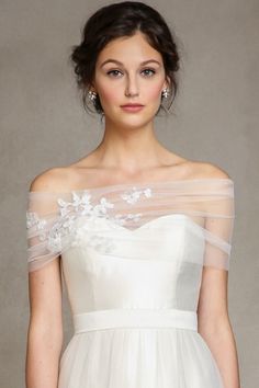 a woman wearing a white dress with sheer sleeves and flowers on the shoulder, standing in front of a gray background