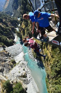 two people hanging from a rope above a river and mountain range with blue water in the foreground