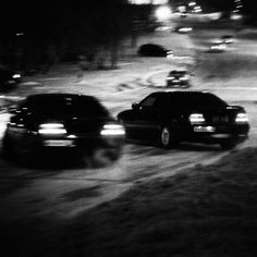 black and white photograph of cars driving down the road at night in snow covered parking lot