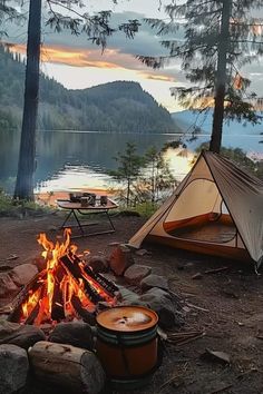 two tents set up next to a campfire on the shore of a lake at sunset