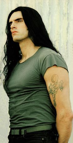 a man with long black hair standing in front of a wall wearing a green shirt